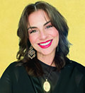 Aixa Pérez-Prado author photo. Aixa has short, wavy brown hair, tan skin, red lipstick, dangly earrings, a gold pendant necklace, and black shirt in front of a yellow background.