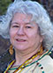 Carla Messinger headshot. Carla has curly gray hair and wears beaded necklaces, earrings, and a green shirt.