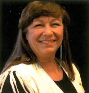 Jeanne Rorex Bridges headshot. Jeanne has long, brown hair with bangs, long earrings, and a white fringe top over a black shirt.