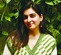 Maham Khwaja headshot: She has long, straight, brown hair and wears a checkered green and white vest in front of a leafy backdrop.